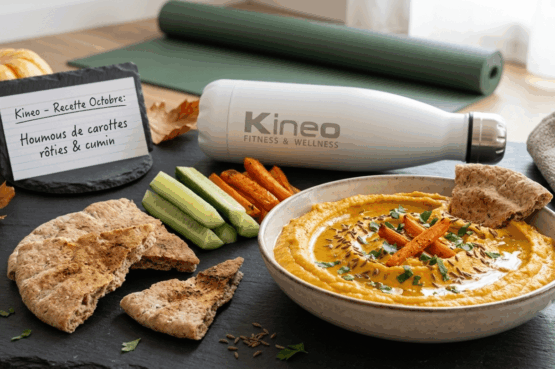 Bol de houmous de carottes rôties au cumin servi avec du pain pita et des bâtonnets de concombre sur ardoise, aux côtés d'une gourde Kineo, d'un tapis de gym et d'un panneau « Kineo - Recette Octobre : Houmous de carottes rôties & cumin »