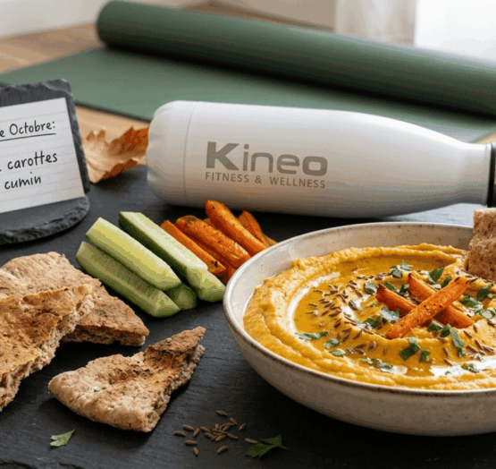 Bol de houmous de carottes rôties au cumin servi avec du pain pita et des bâtonnets de concombre sur ardoise, aux côtés d'une gourde Kineo, d'un tapis de gym et d'un panneau « Kineo - Recette Octobre : Houmous de carottes rôties & cumin »