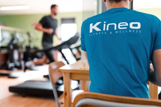 Un homme de dos portant un t-shirt bleu marqué du logo « Kineo Fitness & Wellness », assis à une table dans une salle de sport, avec une personne s'entraînant sur un tapis de course en arrière-plan.