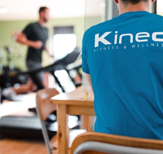 Un homme de dos portant un t-shirt bleu marqué du logo « Kineo Fitness & Wellness », assis à une table dans une salle de sport, avec une personne s'entraînant sur un tapis de course en arrière-plan.